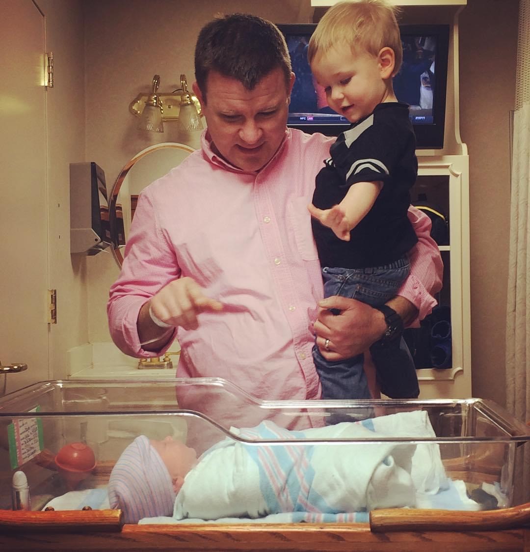 Father in pink shirt introduces young boy to his new baby sister in the hospital