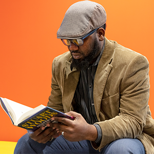Black male with a blazer, hat and glasses on leans forward and looks at a book. 