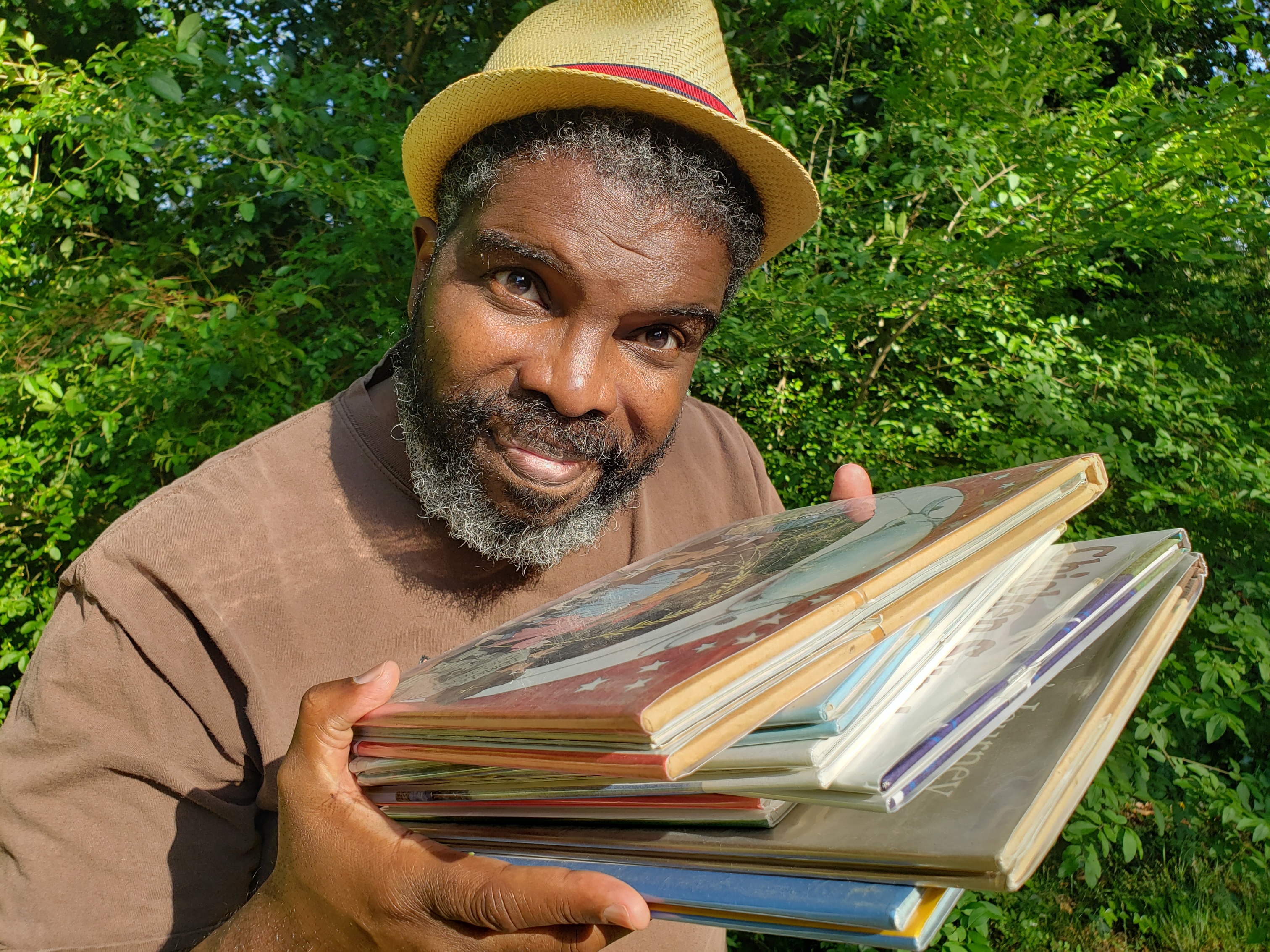 Darion McCloud is a Black man.  He is standing in front of green bushes.  He wears a brown t-shirt and a straw hat.  He is holding a stack of picture books towards the viewer.  His hair and beard are salt and pepper and he shares a slight smile.  