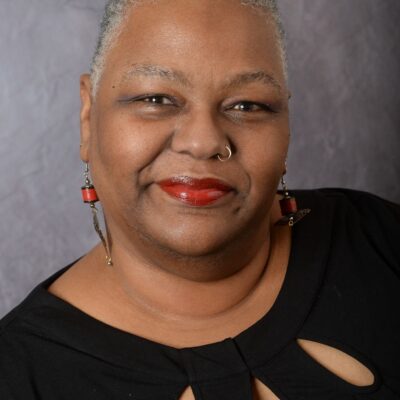 Nancy Tolson is a Black woman.  She is in front of a gray background.  She wears a black top with cut-outs, orangish red lipstick and long beaded earrings.  She also has a nose ring.  Her hair is salt and pepper.  She is giving a slight smile.  