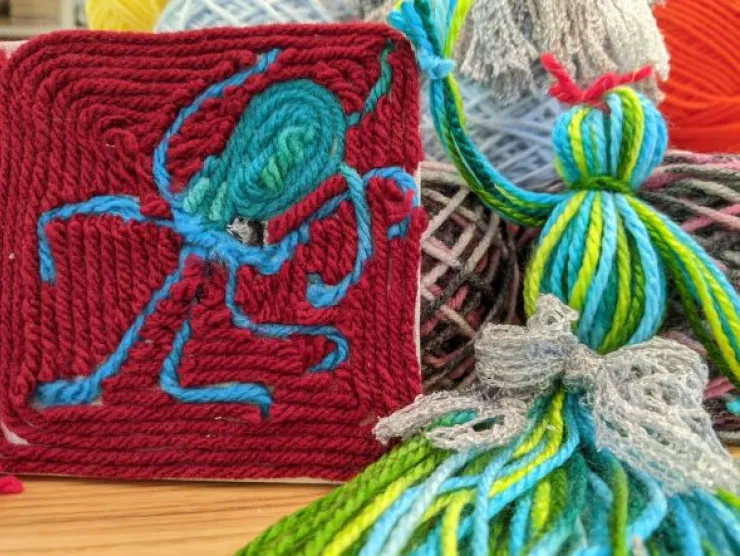 On the left is a yarn painting of an octopus using red yarn for the background and blue green yarn for the octopus. On the right is a yarn doll made with vari-colored green/yellow/blue yarn with a red yarn bow on her head and a silver lacy bow belt.