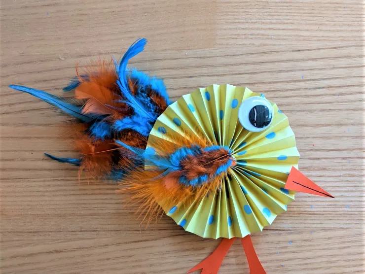 Photo: a bird made from yellow paper with blue polka dots, folded into a circular fan shape with orange and blue feathers for wings and tail, orange paper legs and beak, and a giant googly eye