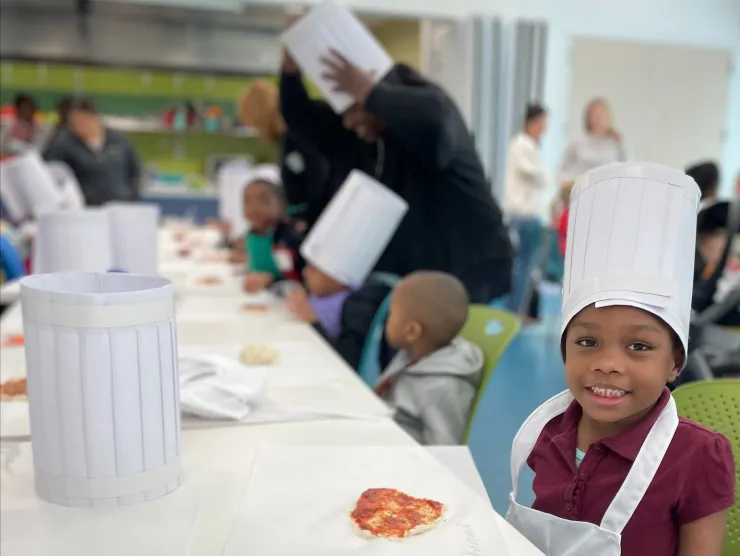 A long table stretches out away from the viewer, with children on both sides of the table wearing chef hats. Small handmade pizzas are on the table in front of each child.