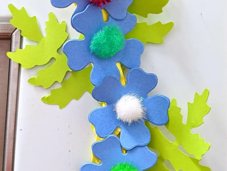 Blue paper flowers with light green leaves with multicolored pompoms as centers - created on a Sizzix machine