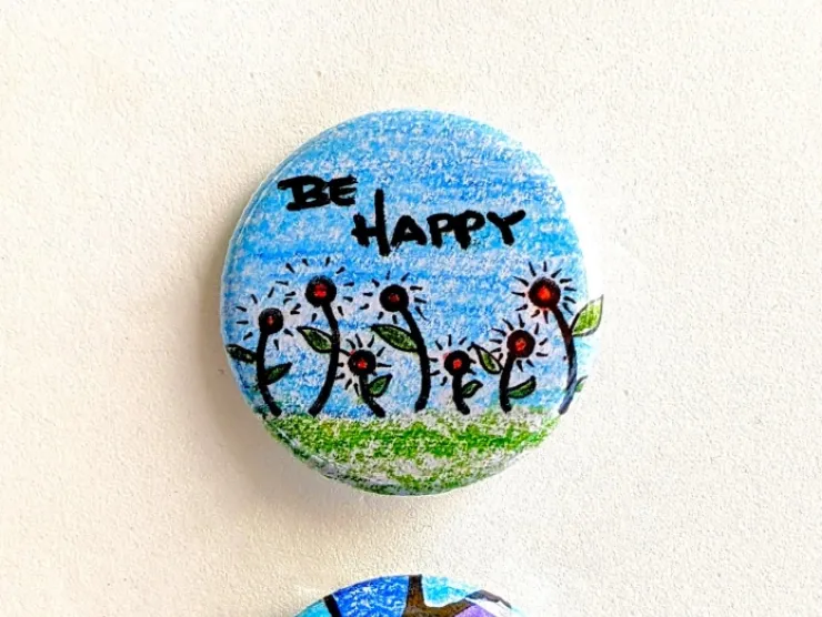 Two pin-back buttons: one has the words "BE HAPPY" written above some hand drawn red flowers on a blue background; the other is a piece of abstract art in blues and purples and black