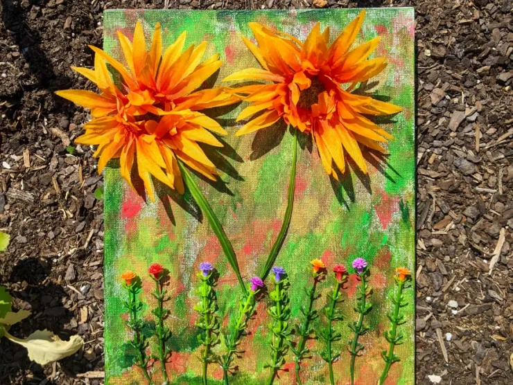 photo: Large orange fake flowers and small multicolored fake flowers mounted on a green/orange/gold/red painted canvas