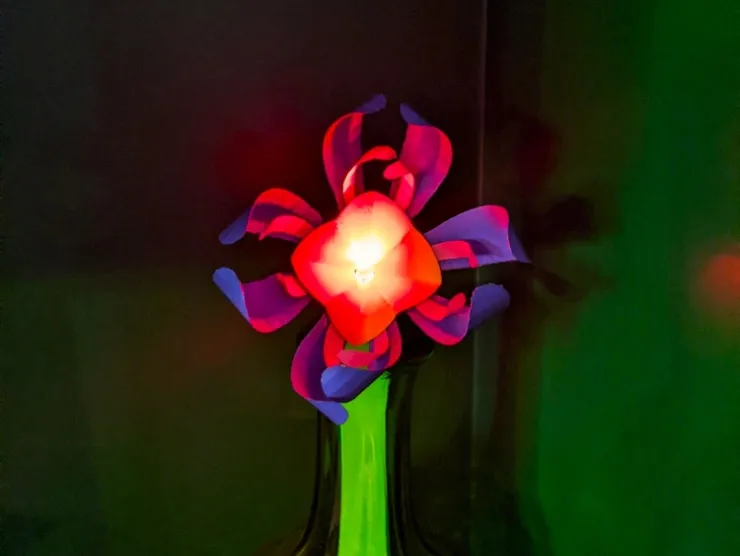 Photo: a paper flower made with purple, blue, and yellow paper petals on a green stem. There is an LED lit up in the middle of the flower.