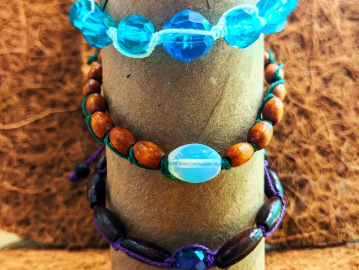 Photo: three bracelets. Blue faceted beads with a light blue cord. Brown wooden beads with a green cord. Dark brown beads with a purple cord.
