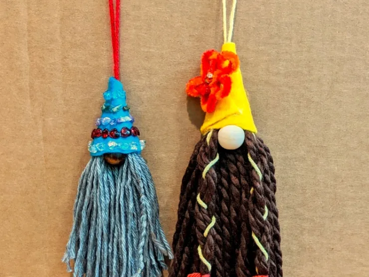 photo: two yarn and felt gnomes, decorated for summer. One has a grey beard, dark brown nose, and a blue hat decorated with stone chips. One has a dark brown beard, light nose, and yellow hat decorated with an orange and red flower. Part of his beard is braided.