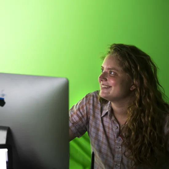 Young white female with long curly hair and a pink shirt sits in front of a MAC computer with a green wall behind her.