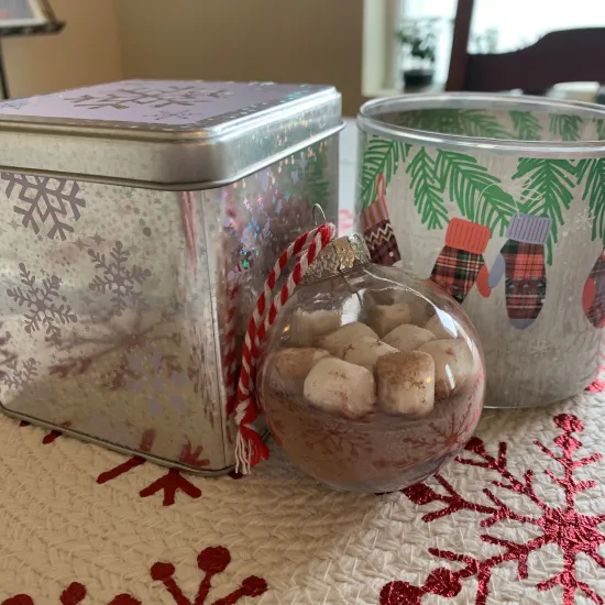 A clear ornament filled with hot cocoa mix and marshmallows, posed against two decorative holiday tins.