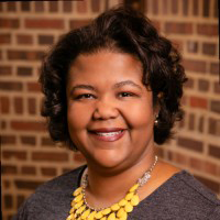 Dr. Nicole A. Cooke is standing in front of a red brick wall and wearing a grey blouse and yellow necklace.