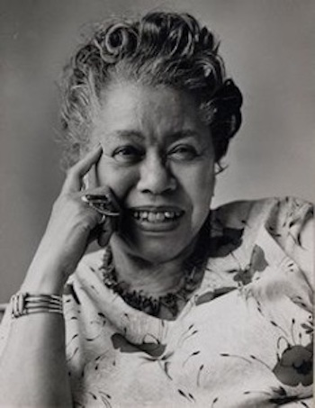 Black and white image of Augusta Baker | An older Black woman wearing a flowered top.  She is smiling with her hand on her chin wearing large silver rings.  
