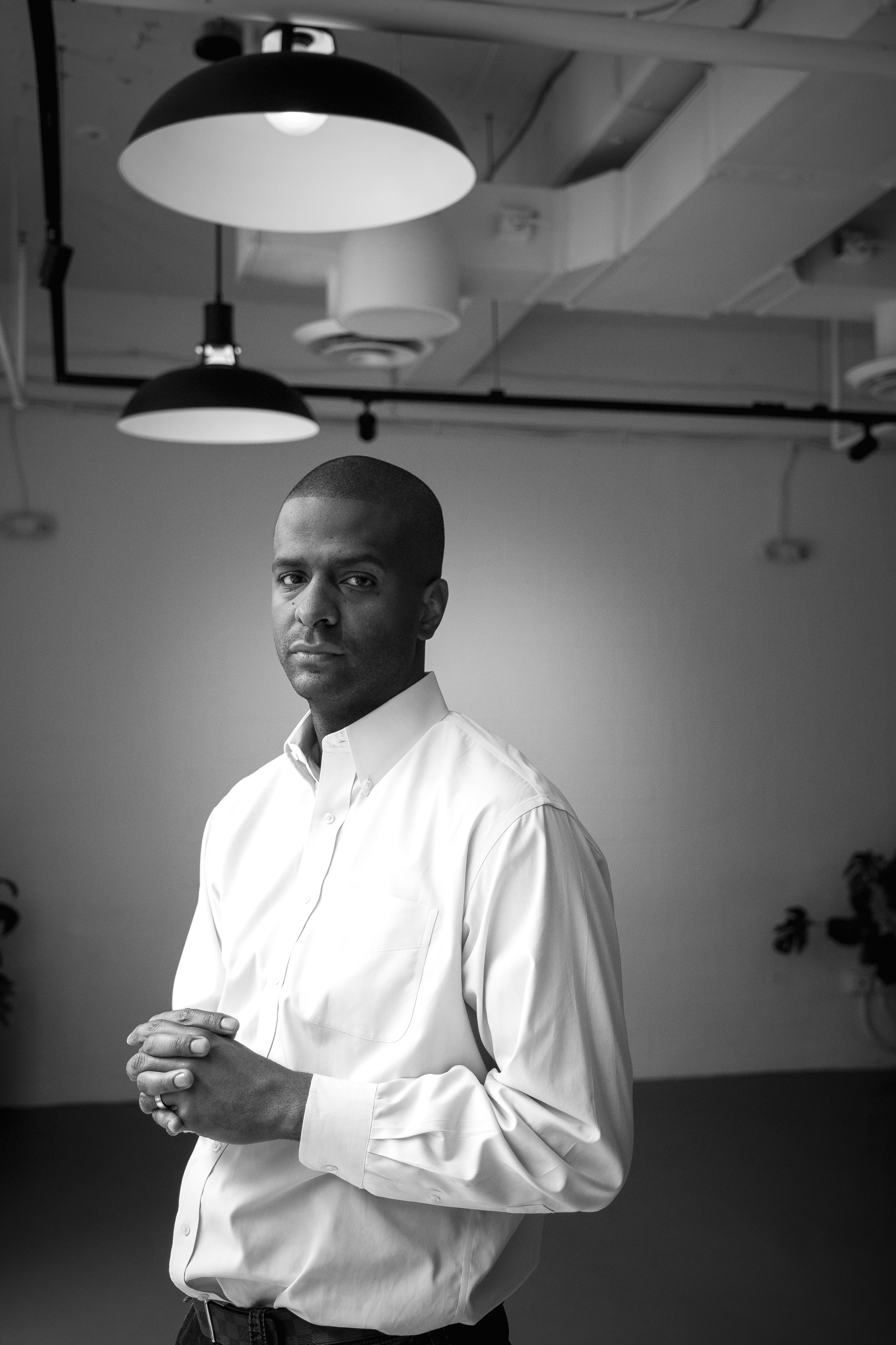 Black and white image of Bakari Sellers | an adult Black man in a white button-down shirt with his hands clasped in front of him.  He looks serious.  Two large light fixtures are above him.  