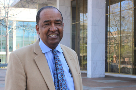 An older African American man in a tan suit, blue shirt and tie smiling and standing in front of a building with windows.