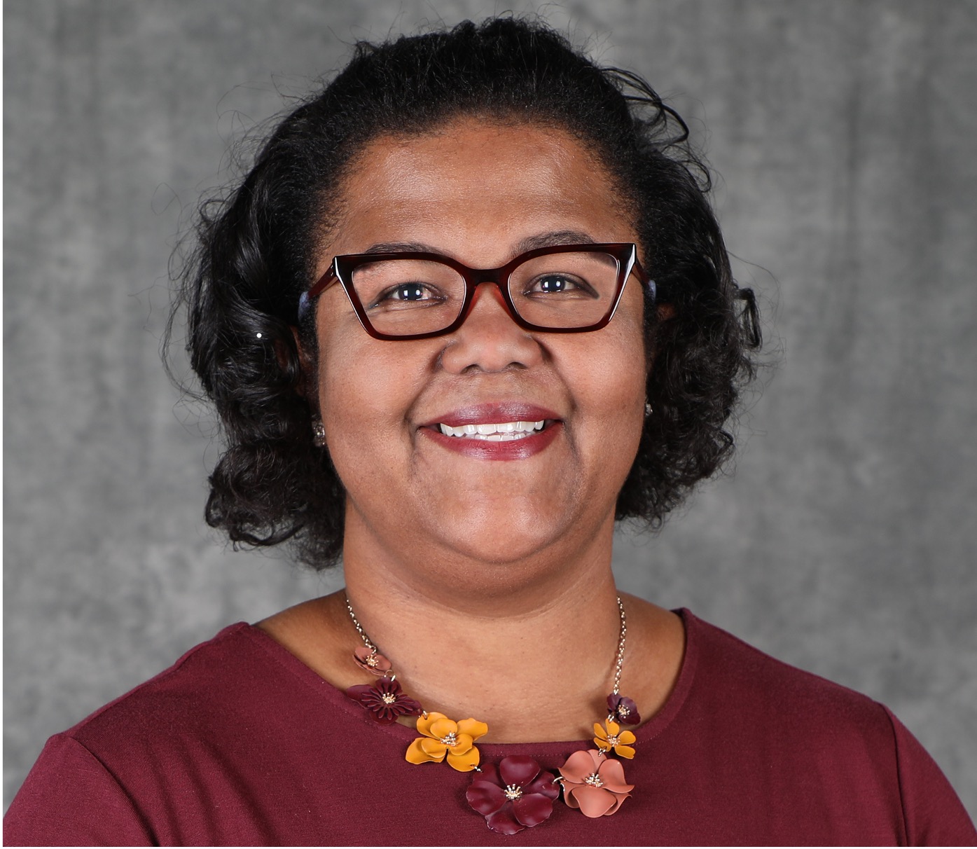 Photo of Dr. Nicole Cooke. | She is a Black woman wearing maroon-colored glasses.  She also has on a maroon-colored top and a necklace with maroon-colored flowers.  