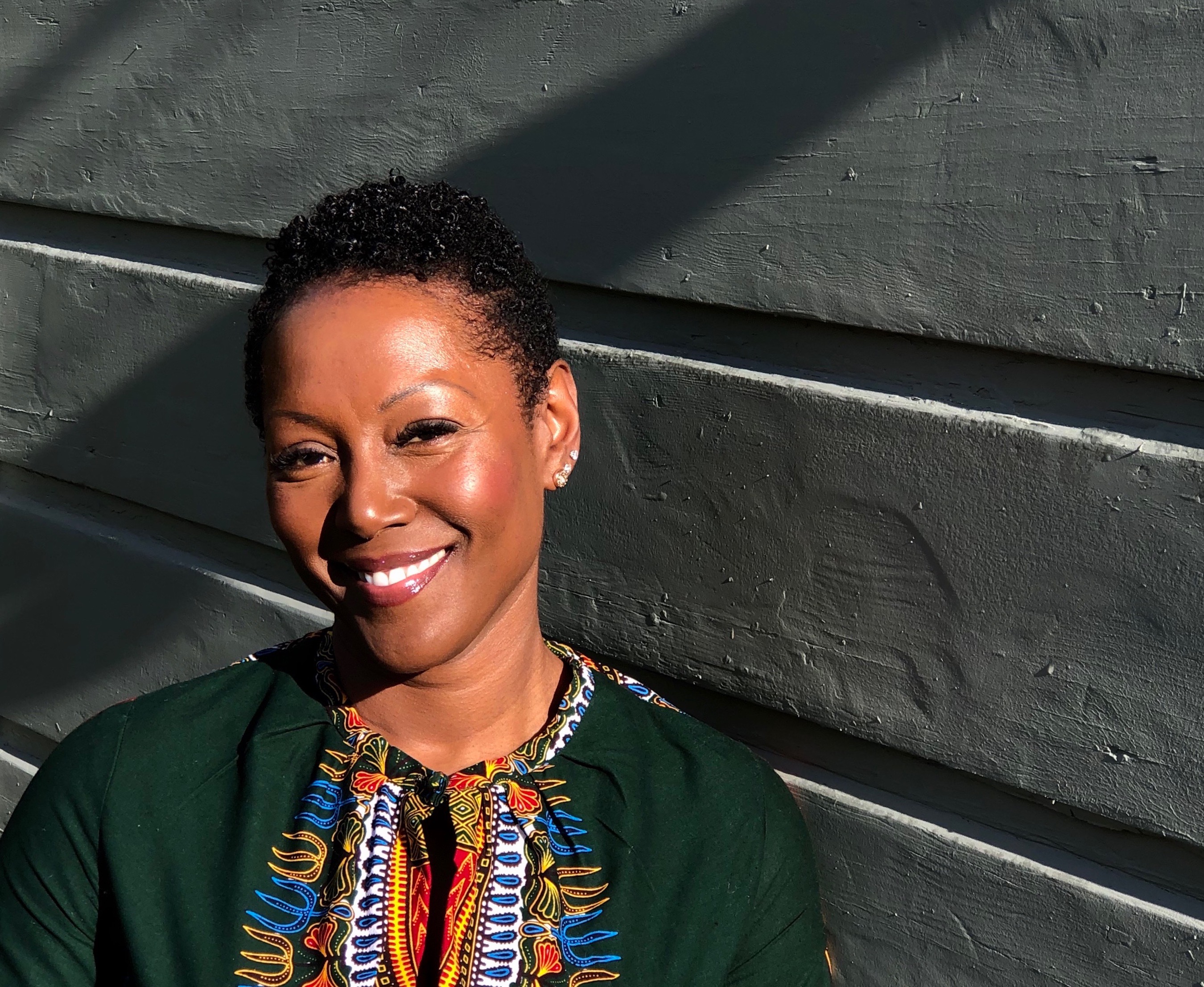Photo of Dr. Monique Morris | A Black woman photographed against a building.  She is wearing a dark green top with an African print.  The sunlight is streaming down and she is smiling.  