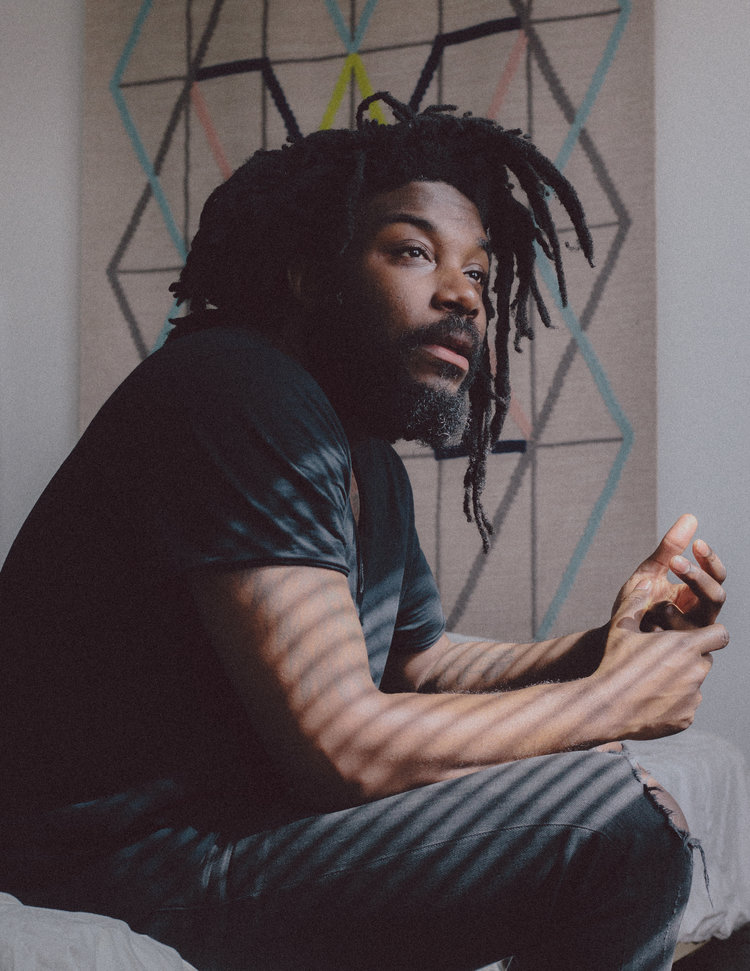 Photo of author Jason Reynolds | Black Man wearing a black t-shirt and jeans sitting and staring in the distance.  He is shadowed by window blinds and is sitting in front of a graphic art image.  