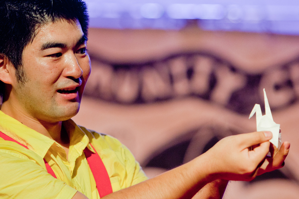 Image of Yasu Ishida, an Asian man wearing a yellow shirt with red suspenders.  He is holding a white paper crane and smiling.