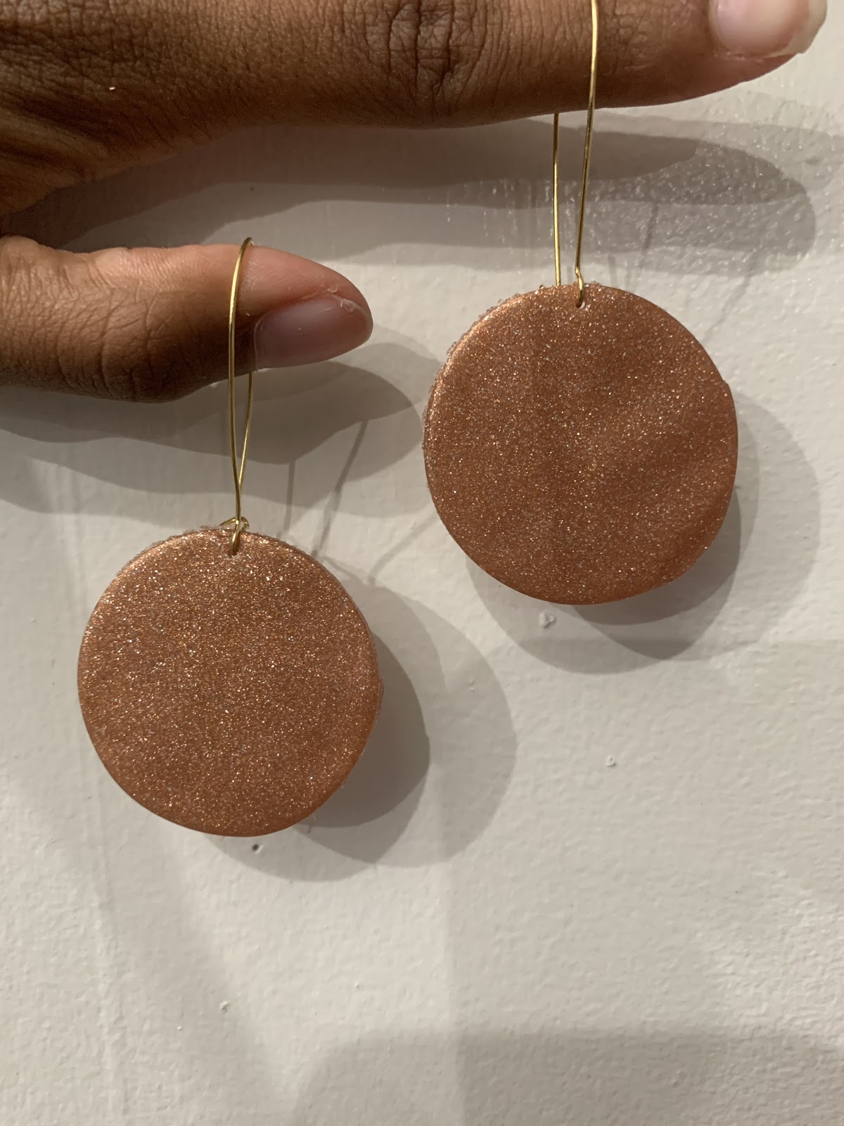 Bronze circular clay earrings with hanging thin wires hanging on two brown skinned fingers, against a white background.
