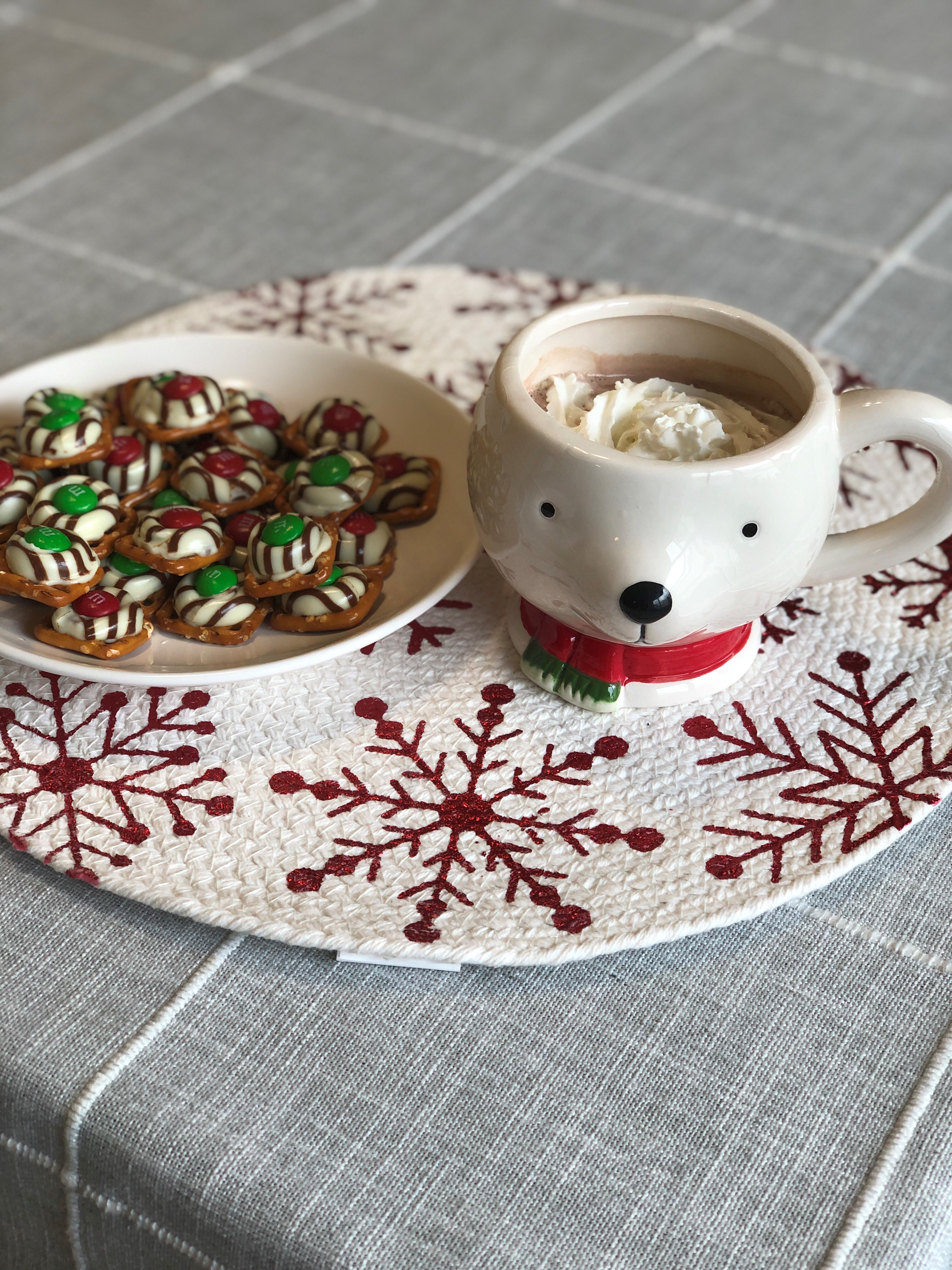 Picture of a ceramic mug shaped like a polar bear head wearing a red scarf around its neck, next to a plate or pretzel hug candies. The mug is filled will hot chocolate and whipped cream. The mug an plate sit on a white quilted placemat white a red snowflake pattern.