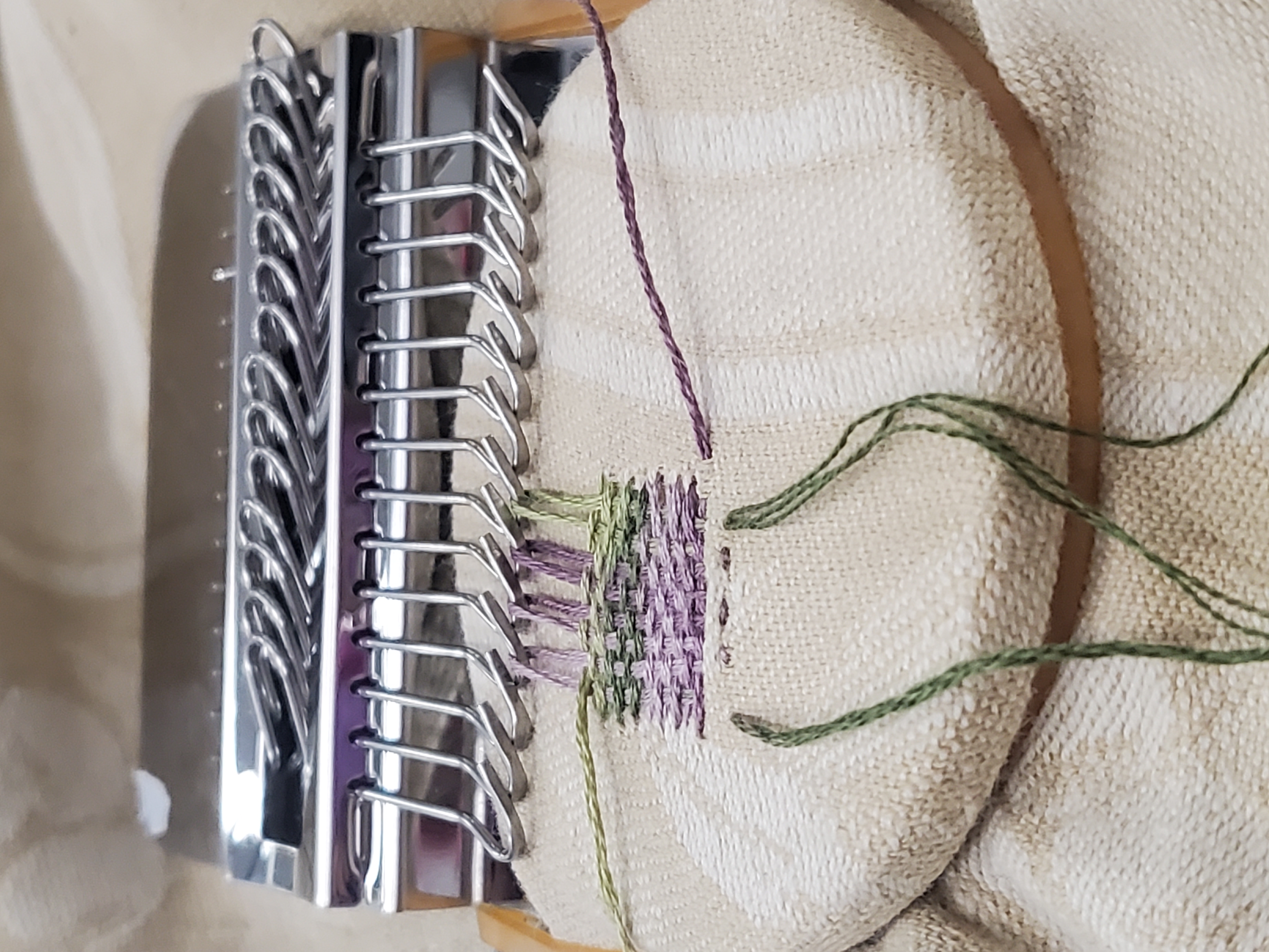 metal darning loom holds green and purple stitches on cream fabric