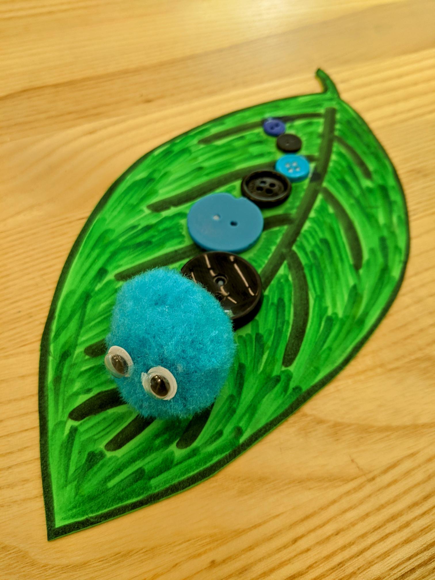 Caterpillar made from blue and black buttons in graduated sizes with a blue pompom head, sitting on a green foam leaf