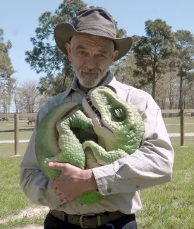 Image of Mr. Ed, an older white man.  He wears a hat and safari-style clothing.  In his arms, he cradles a dinosaur puppet.