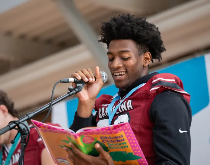 Image of USC Gamecock Football Player reading Chicka Chicka Boom Boom at a microphone.  The player is a young Black man with short twists.  He wears a garnet jersey with black sleeves.  He smiles as he holds the mic reading the book.  
