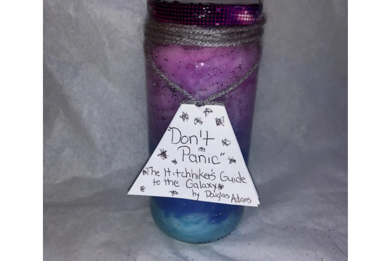 Mason Jar with different color cotton (pink, purple, periwinkle, and blue) and glitter (silver and purple) inside. A note hangs on grey yarn and says "Don't Panic" The Hitchhikers Guide to the Galaxy by Douglas Adams