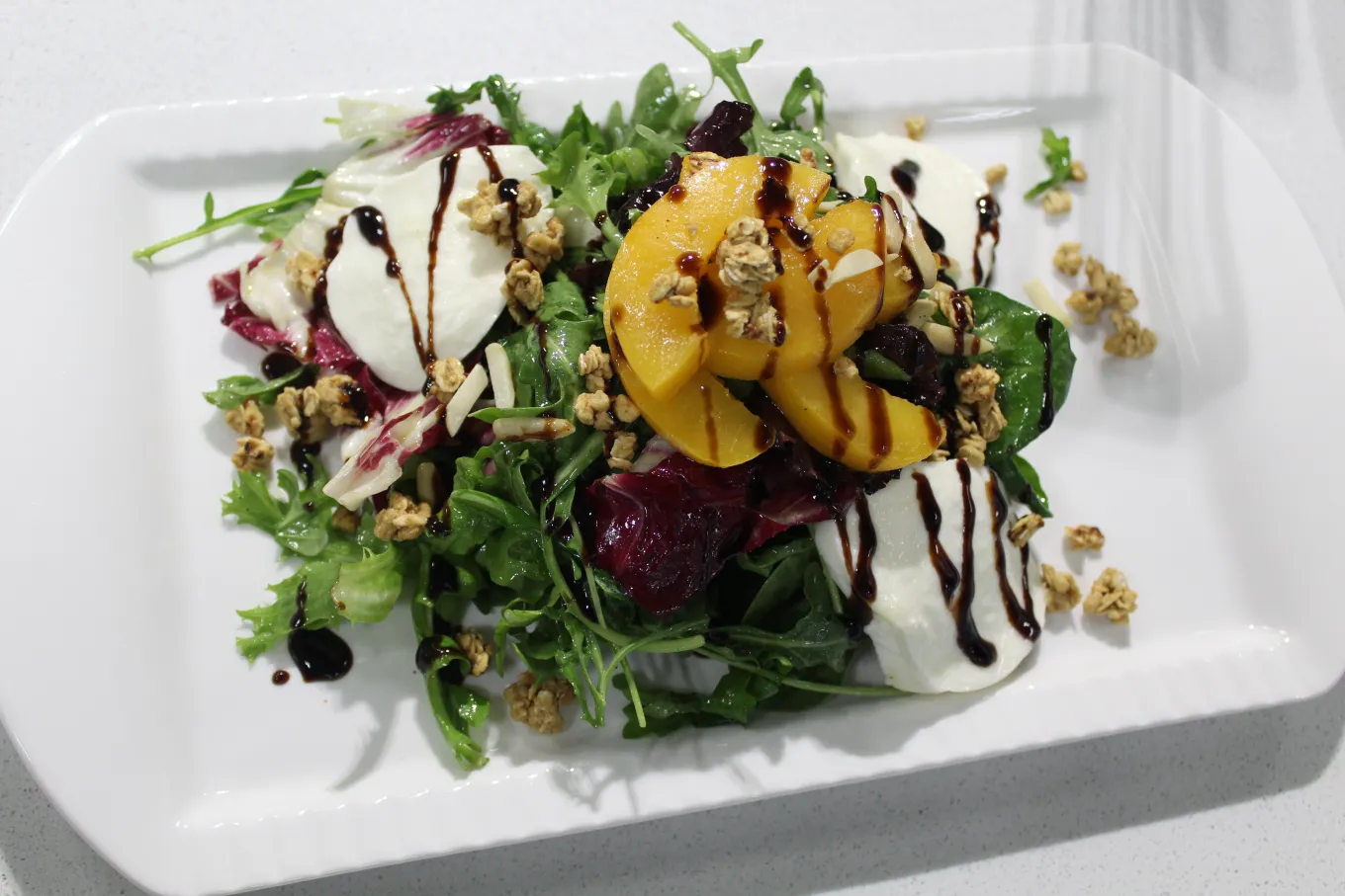 Colorful salad topped with peaches, mozzarella, greens, and balsamic drizzle.