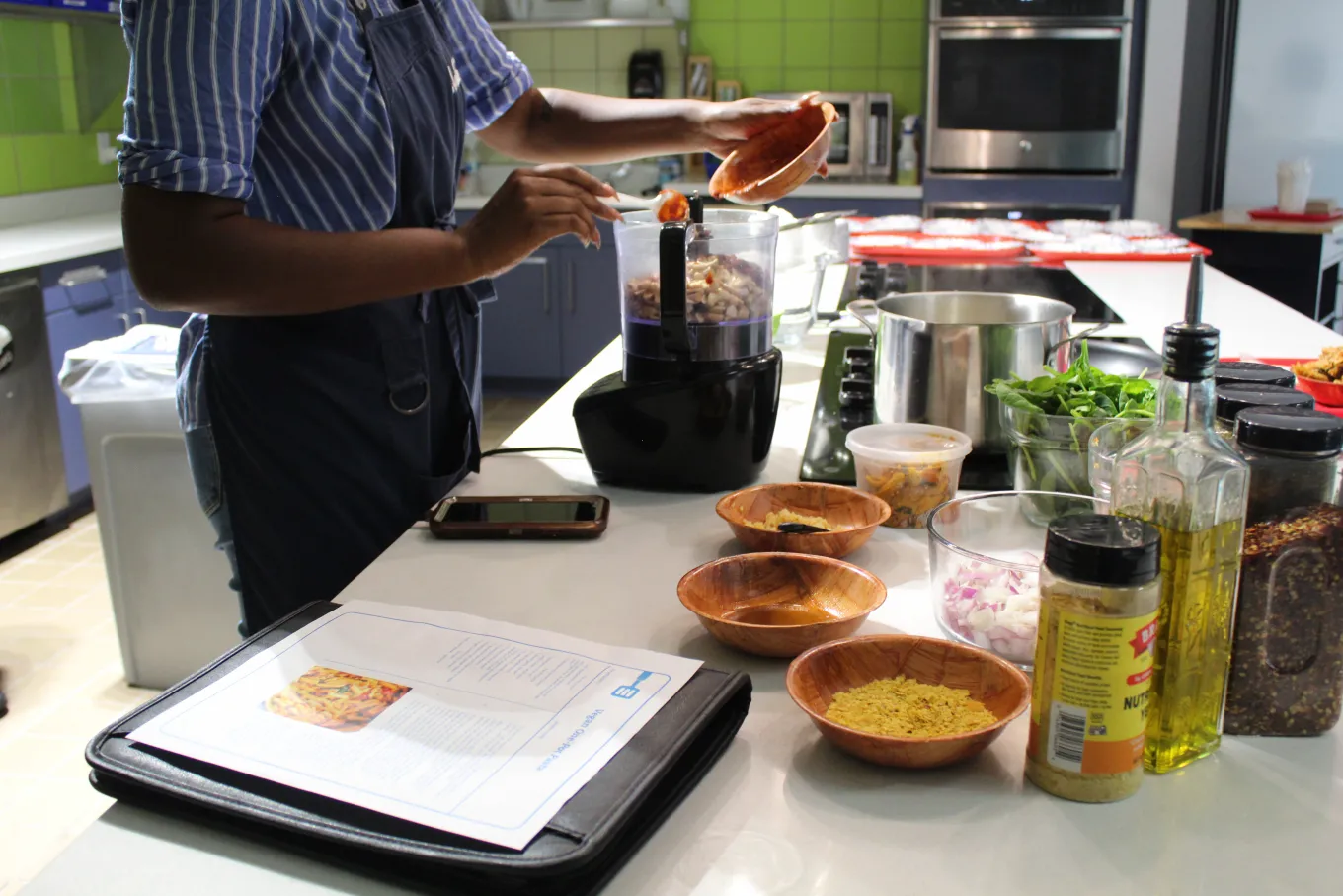 Participant adding ingredients to a food processor with spices and vegetables laid out on the counter.