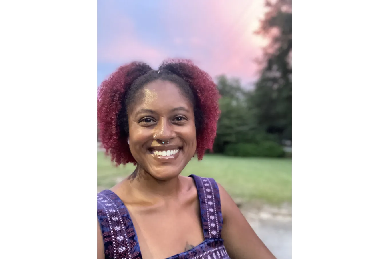 selfie of a smiling person wih curly hair, dark roots and dark red dyed hair. the background is nature with a sunset