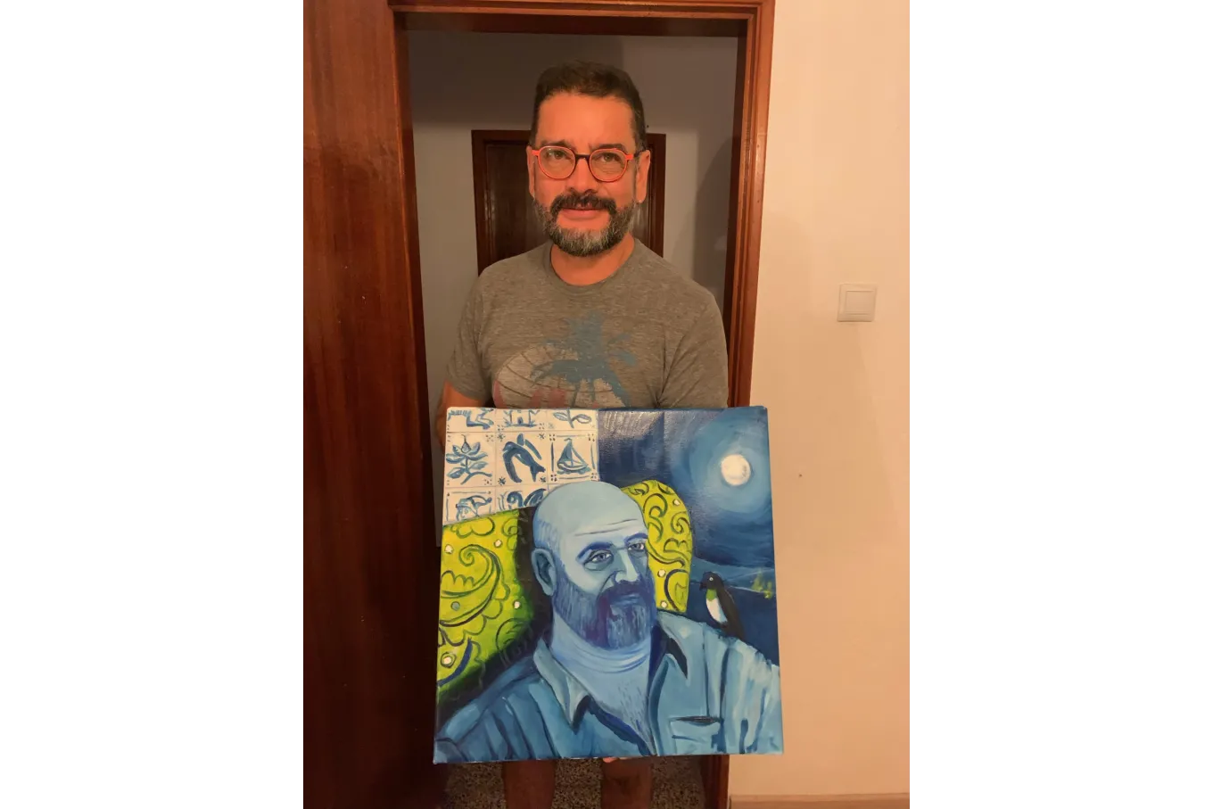 Garcia Lemos, A. Standing in a doorway, holding a portrait he has painted. Clothed in a graphic tshirt and glasses