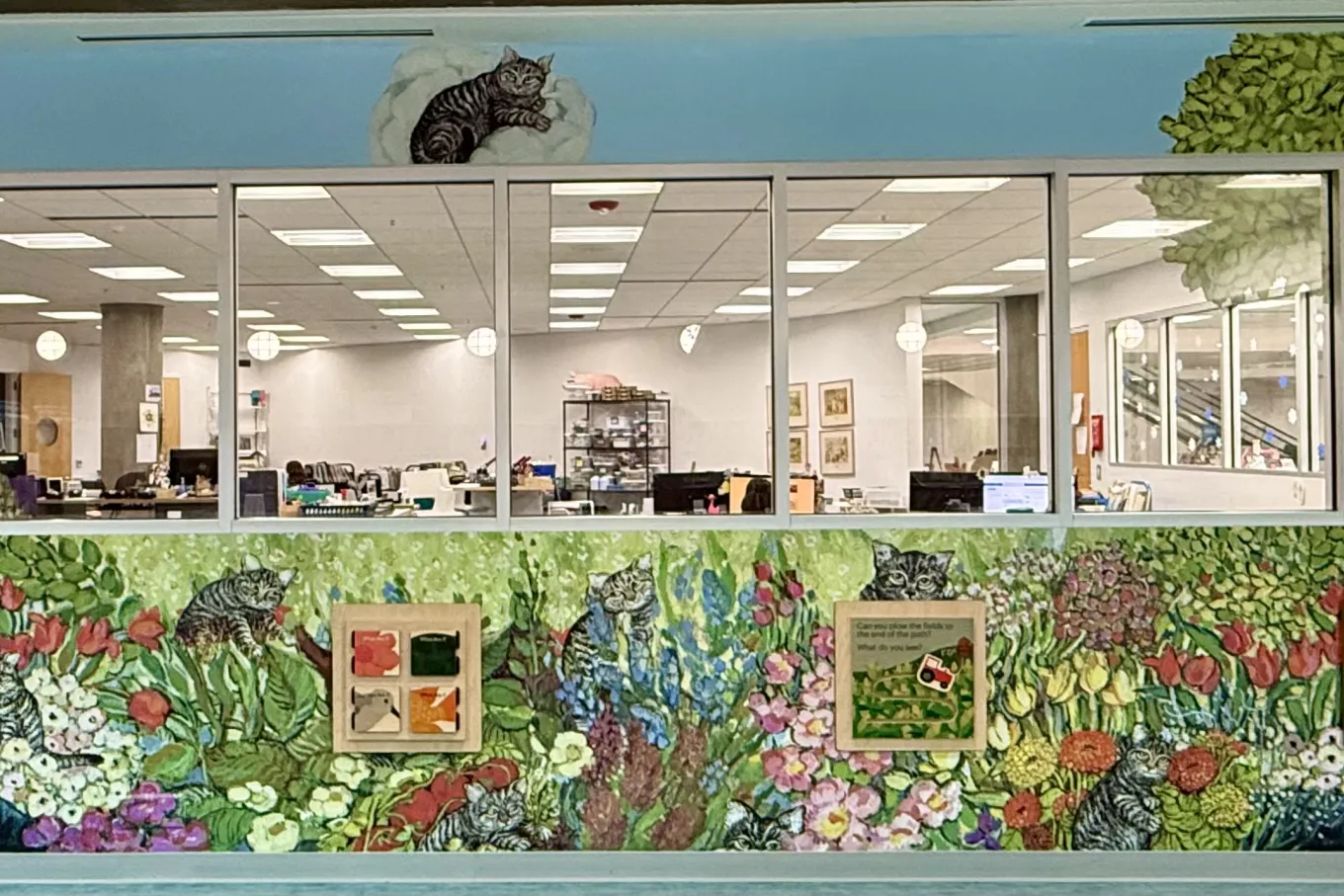 A colorful wall mural surrounds a row of office windows, depicting a lush garden filled with bright pink, purple, yellow, and white flowers. Gray tabby cats hide among the plants and lounge atop the window frames. A red house with a steep roof sits on the left, while a large leafy tree frames the right side. Through the windows, desks, computers, and fluorescent lights are visible. Small framed interactive panels line the lower part of the mural.