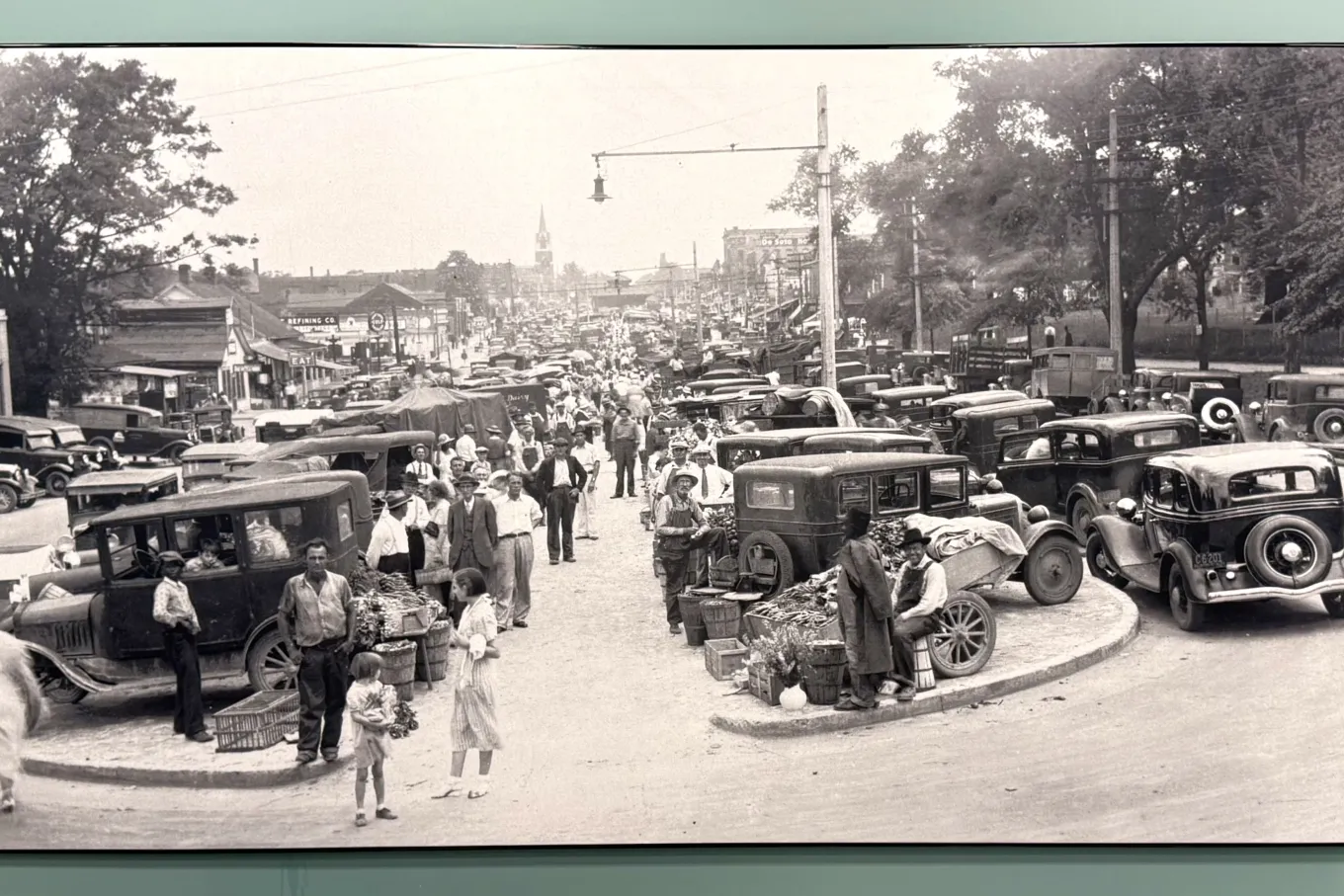 A large black-and-white historic photo shows a busy street market from the early automobile era. Vintage cars are parked bumper to bumper along both sides and down the center of a wide street. Vendors sell produce from wooden crates and baskets on sidewalks and small medians. Well-dressed men, women, and children fill the scene. In the foreground, a child stands by stacked vegetable crates. Storefronts and utility poles lead toward a distant church steeple, capturing a lively small-town marketplace.