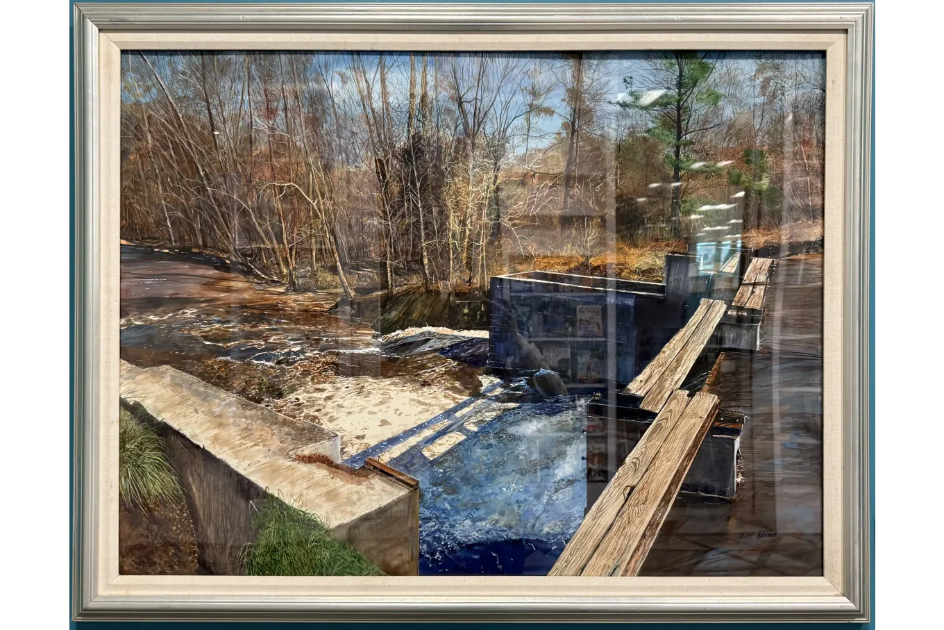 A framed landscape painting shows a small dam along a wooded river in late autumn or early winter. Brown water spills over a low concrete spillway, forming white foam as it falls into a deep blue pool. A narrow wooden walkway runs beside metal gates that control the flow. Bare trees and a few evergreens stand in the background, reflected in the cool, quiet water.