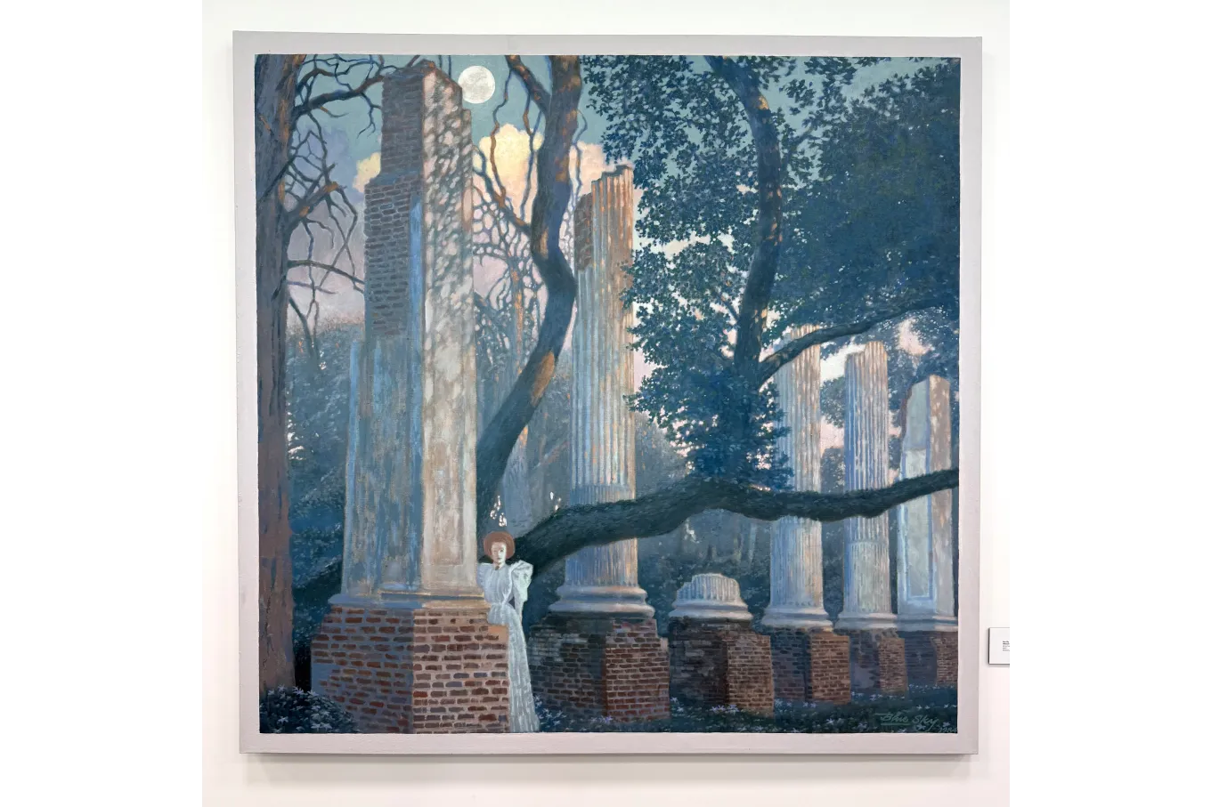 A large painting depicts a dreamlike nighttime scene of classical ruins in a wooded landscape. Tall, weathered white columns rise from brick bases, some broken and crumbling. A dark, twisting tree stretches across the foreground. A full moon glows in a blue sky, casting soft light. Near one column, a young woman with short reddish hair in a long pale dress leans against a brick pedestal, calm and contemplative in the cool, mysterious scene.