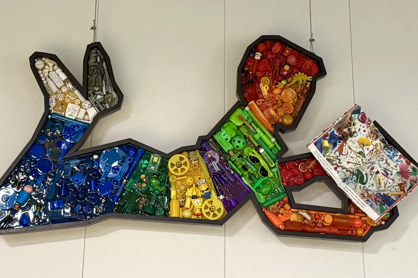 A wall-mounted mixed-media artwork shows the silhouette of a child reading while lying on their stomach with legs bent upward. Outlined in black, the figure is divided into sections filled with small plastic objects arranged in a rainbow gradient—reds and oranges at the head and hands, shifting through yellow, green, blue, and purple, with white at the feet. Hanging by thin wires, the piece blends playful imagery with colorful recycled materials.