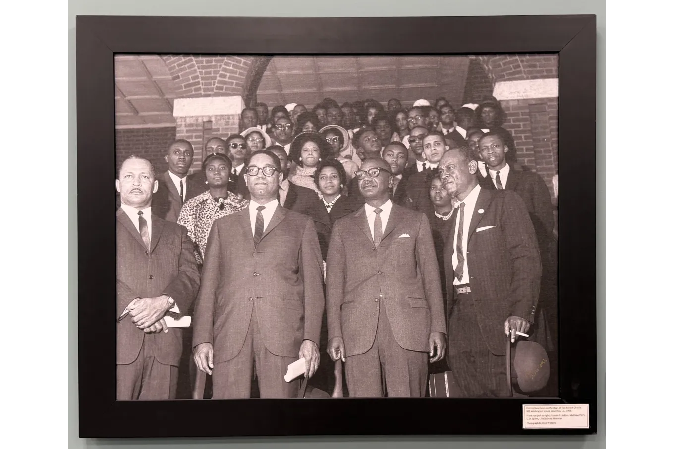 A framed black-and-white photograph shows a large group of African American men and women standing closely together on brick steps beneath an arched entryway. Four middle-aged men in suits and ties stand in front, facing forward with serious expressions. Behind them, formally dressed adults fill the steps outside a public building, conveying a sense of unity and purpose.