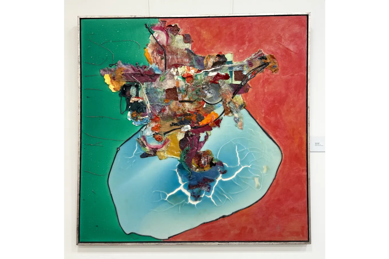 A large framed abstract mixed-media artwork features a vertically split background—deep green on the left and vivid red on the right. At the center, a raised textured cluster of layered paint, resin, and collage elements stands out. Below it, an irregular glossy pale blue shape, outlined in black with white vein-like cracks, contrasts sharply with the flat, bold color fields and sculptural surface details.
