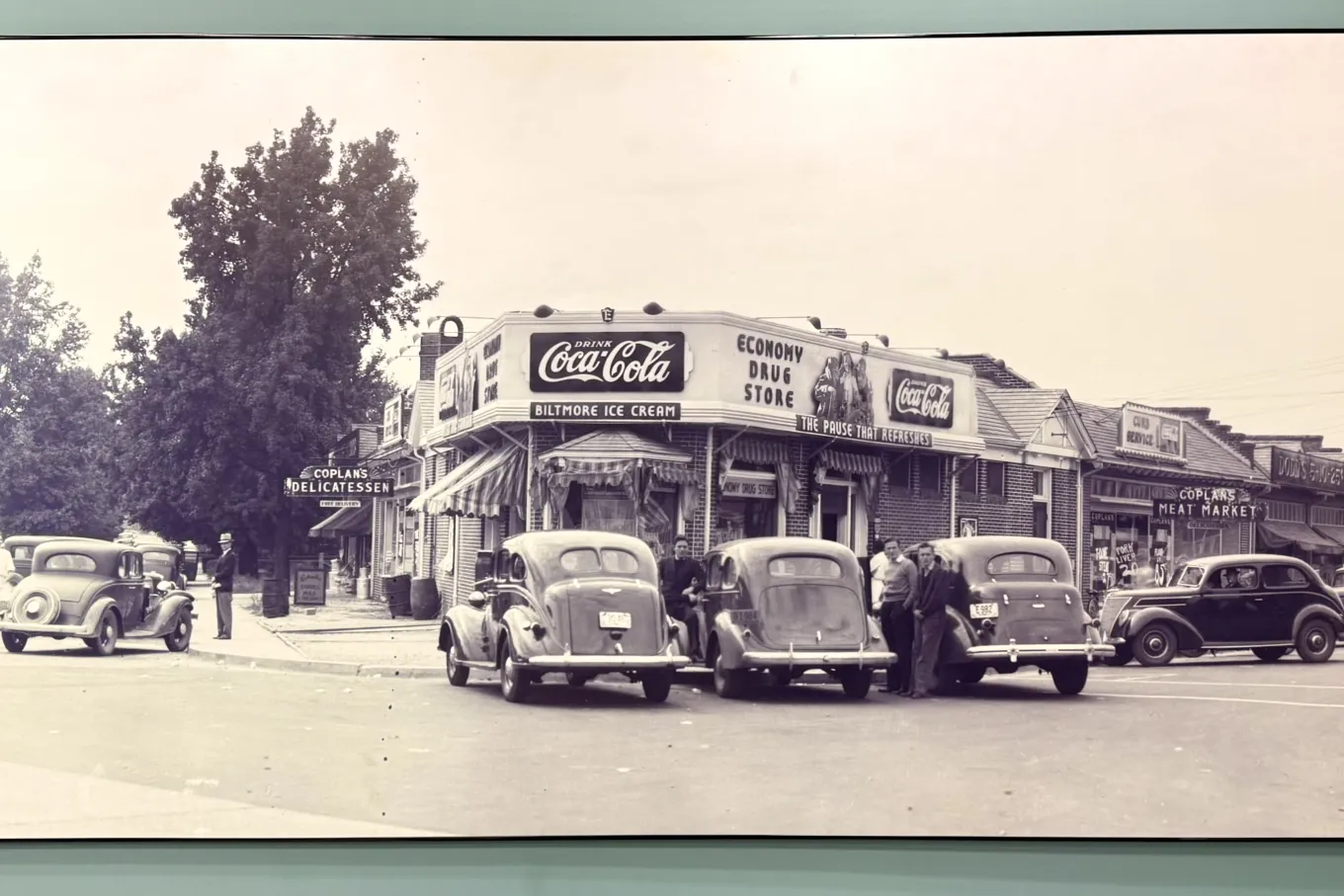 A framed black-and-white photo shows a 1930s street corner with small brick storefronts. An Economy Drug Store features prominent Coca-Cola signs and a Biltmore Ice Cream awning. Vintage cars line the curb as a few men stand nearby talking. Signs for a delicatessen and meat market appear along the row of shops, and a large tree rises behind the buildings, capturing a lively neighborhood scene from the early automobile era.