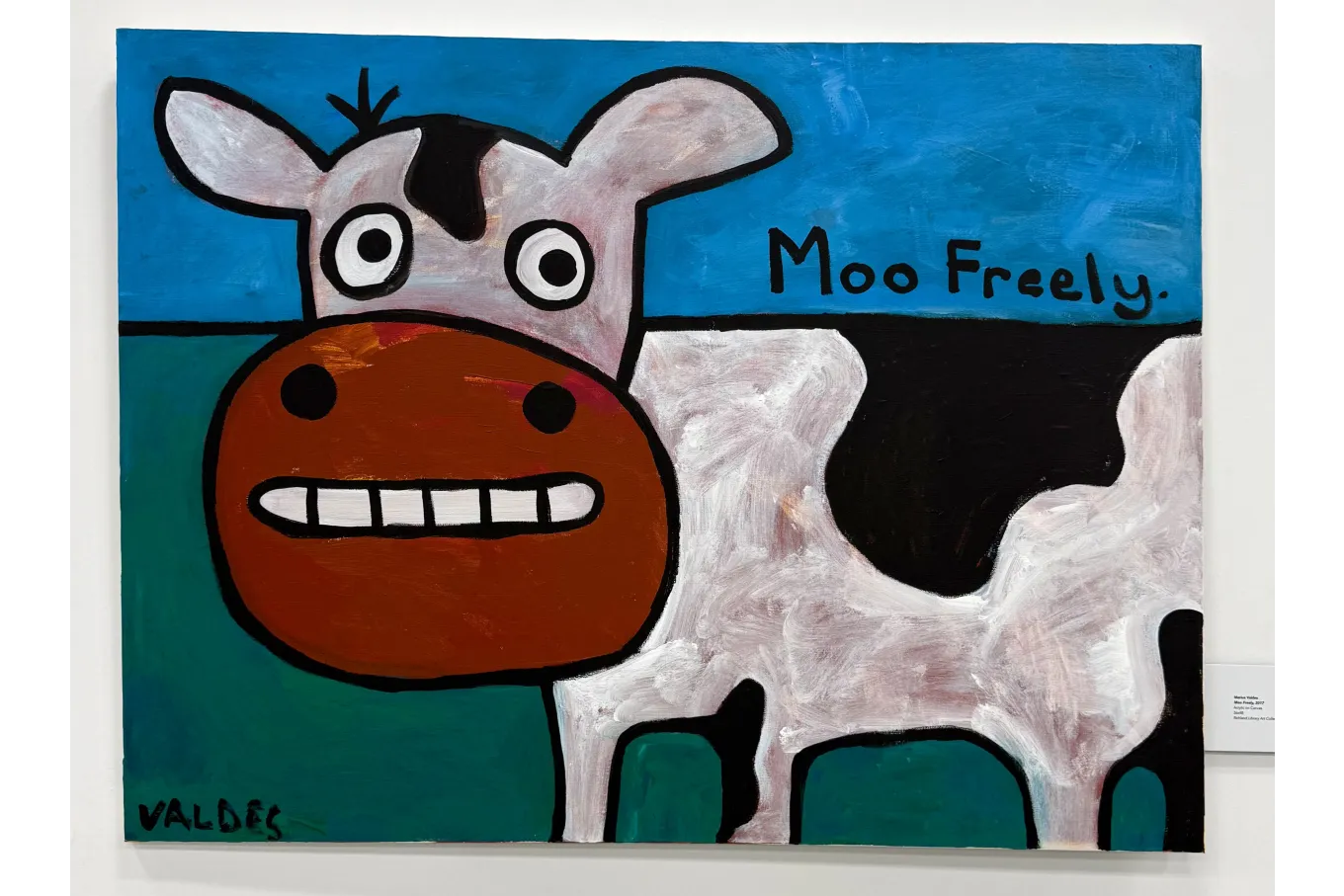 A large, brightly colored painting shows a cartoon-style cow with wide, round eyes and a big red-orange muzzle standing against a simple blue sky and green ground. The cow’s body is white with bold black patches outlined in thick black lines, and the words “Moo Freely.” appear in black lettering to the right. The playful, simplified style and vivid colors give the artwork a cheerful, lighthearted feel.