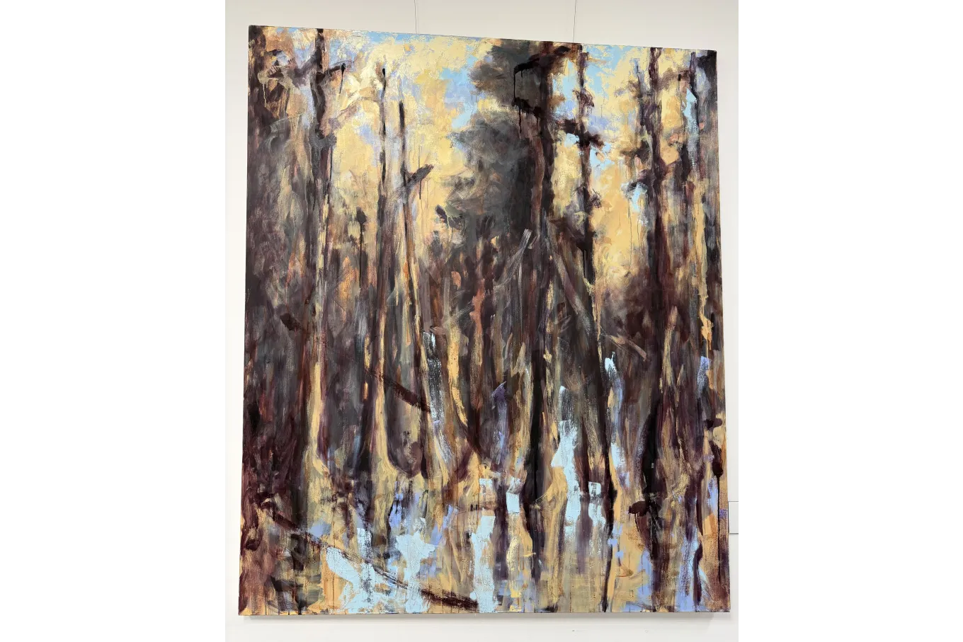 A large abstract painting suggests a forest scene through expressive vertical brushstrokes in deep browns, muted purples, and smoky grays rising against a warm golden-yellow and pale blue background. The tall, tree-like forms appear layered and partially blurred, with drips and loose marks creating a sense of movement and atmosphere, evoking light filtering through a dense woodland.