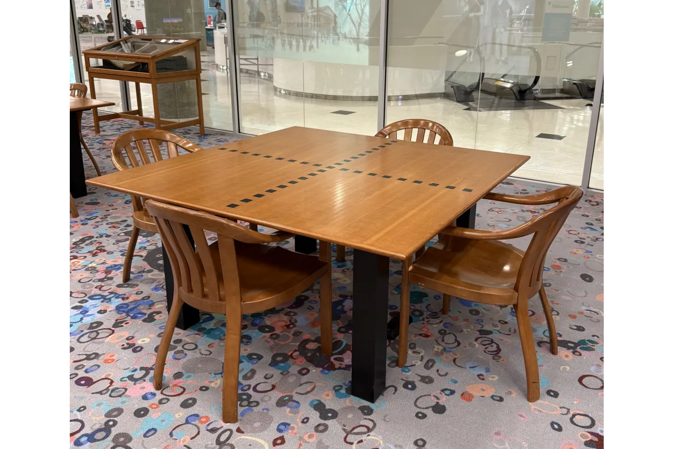 A wooden square table with a smooth, light-brown surface sits in a library space, supported by sturdy black legs and surrounded by four matching wooden chairs with curved backs. Small black square inlays form a cross pattern across the tabletop. The table stands on a colorful patterned carpet, with glass walls and display cases visible in the background.
