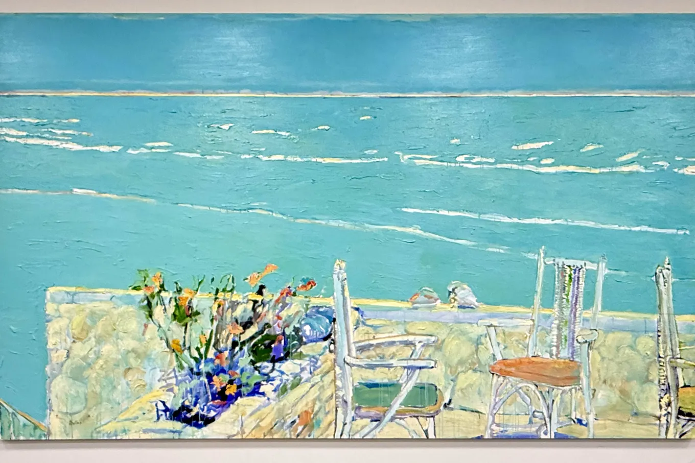 A wide, impressionistic painting depicts a seaside view from a terrace overlooking a calm turquoise ocean. In the foreground, a stone wall borders a small patio with light-colored chairs and a planter filled with bright flowers, while gentle white waves stretch across the horizon beneath a clear blue sky.