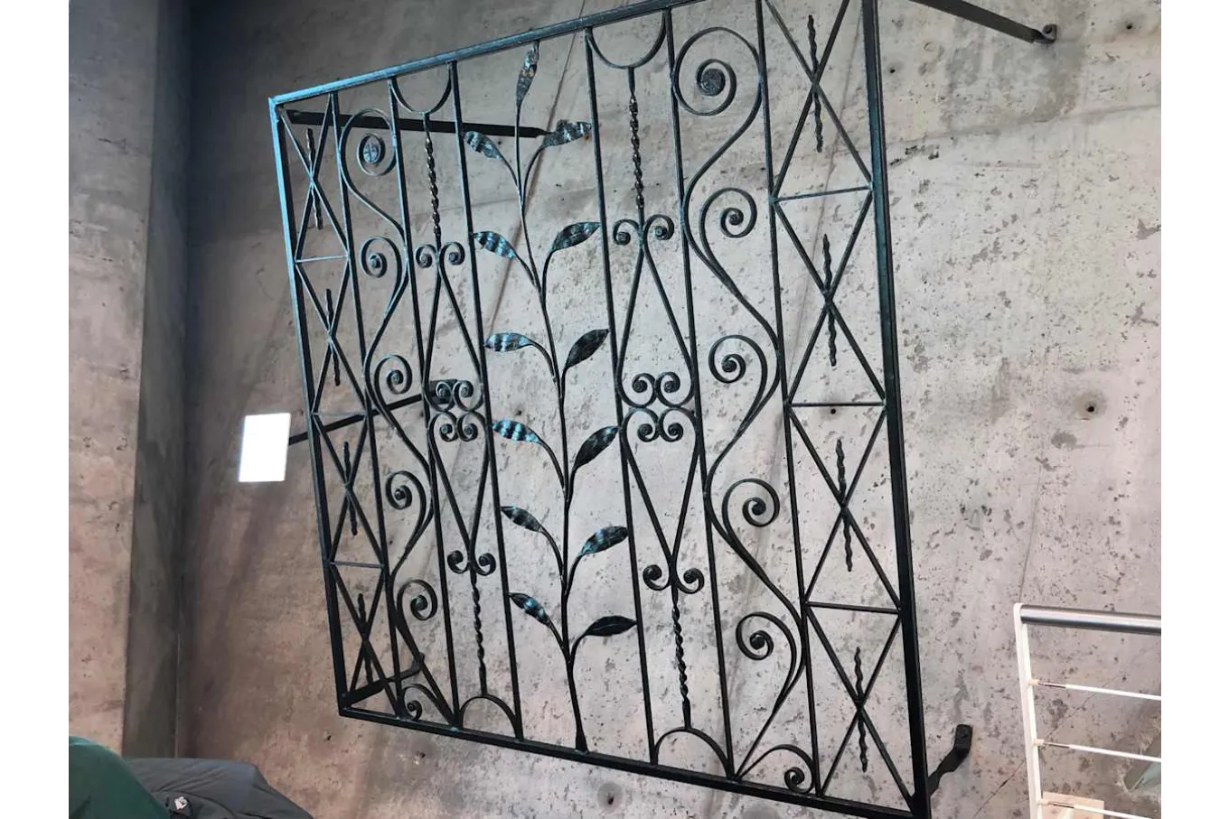 A decorative wrought-iron gate panel is mounted against a concrete wall, featuring symmetrical scrollwork, vertical bars, and geometric patterns. At the center, a stylized vine with metal leaves rises upward, adding an organic element to the structured design. The dark metal contrasts with the light concrete background, highlighting the craftsmanship and ornamental detail.