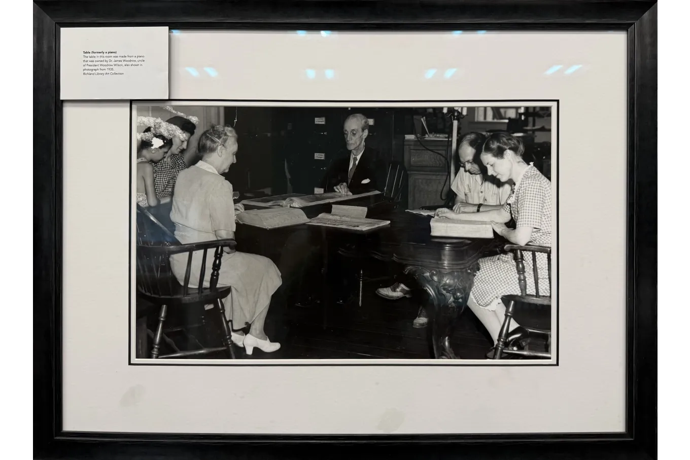 A framed black-and-white photograph shows a small group of adults seated around a large wooden table, reading oversized books or ledgers in what appears to be a library or archival room. An older man sits at the head of the table while others—men and women dressed in mid-20th-century attire—study the open volumes attentively. A caption in the frame identifies the table as formerly a piano, adding historical context to the scene.