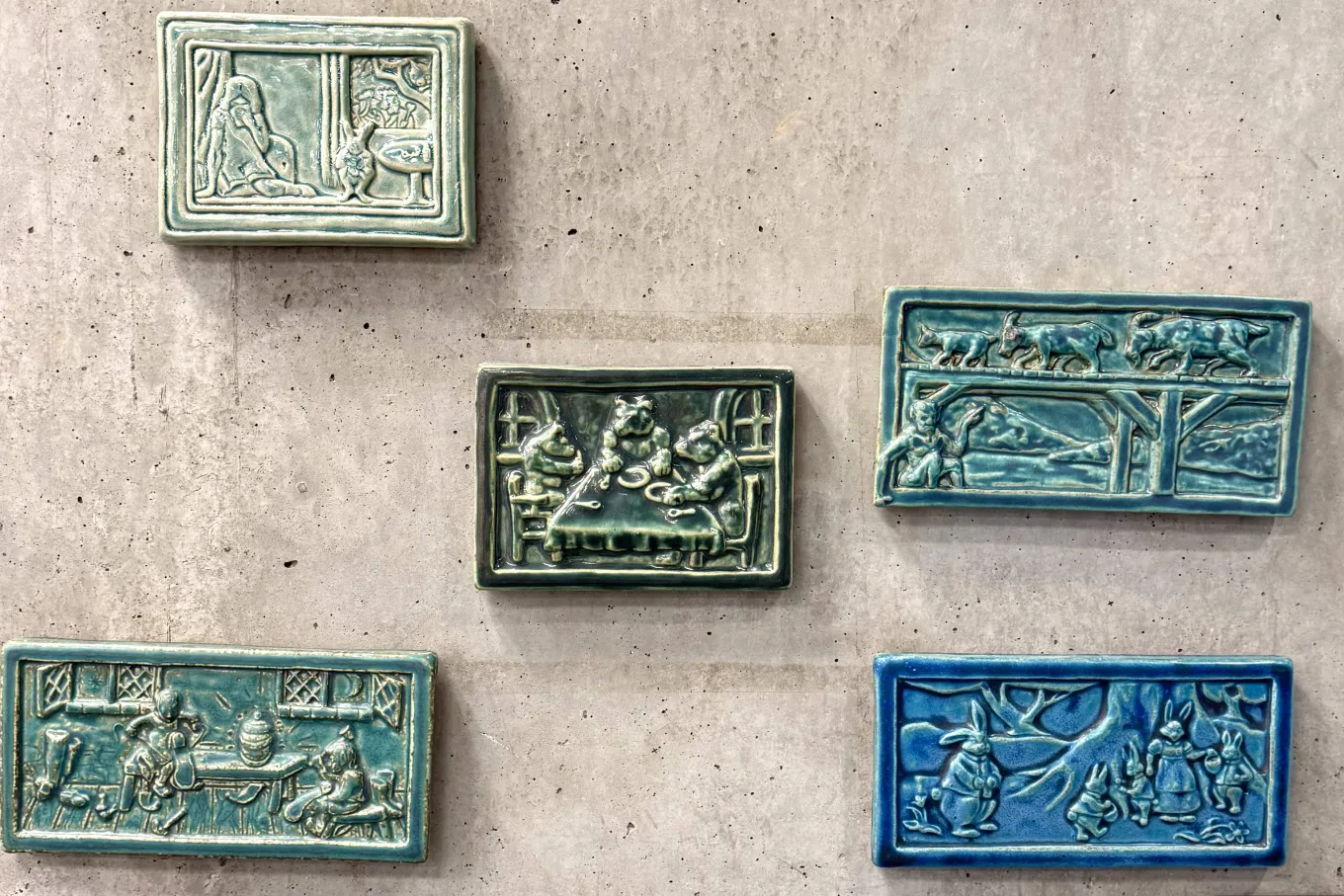 Five small rectangular ceramic tiles in shades of teal and blue are mounted on a concrete wall, each featuring a raised, storybook-style scene in relief. The tiles depict animals and figures in domestic and woodland settings—such as creatures gathered around a table, rabbits beneath a tree, and animals walking along a bridge—rendered with detailed textures and framed borders. The glossy glaze and sculpted surfaces give the pieces a tactile, illustrative quality reminiscent of classic children’s tales.