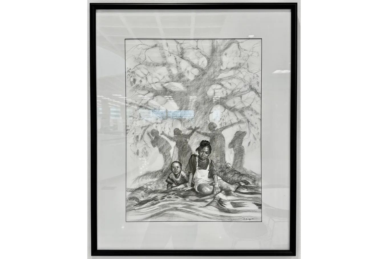 A framed black-and-white drawing shows two children seated beneath a large, spreading tree with textured bark and broad leaves. In the foreground, a young girl sits facing forward with a thoughtful expression while a younger child rests beside her, and open books lie on the ground. Behind them, shadowy silhouettes of other figures reach upward into the branches, creating a layered, symbolic scene that suggests themes of learning, community, and growth.